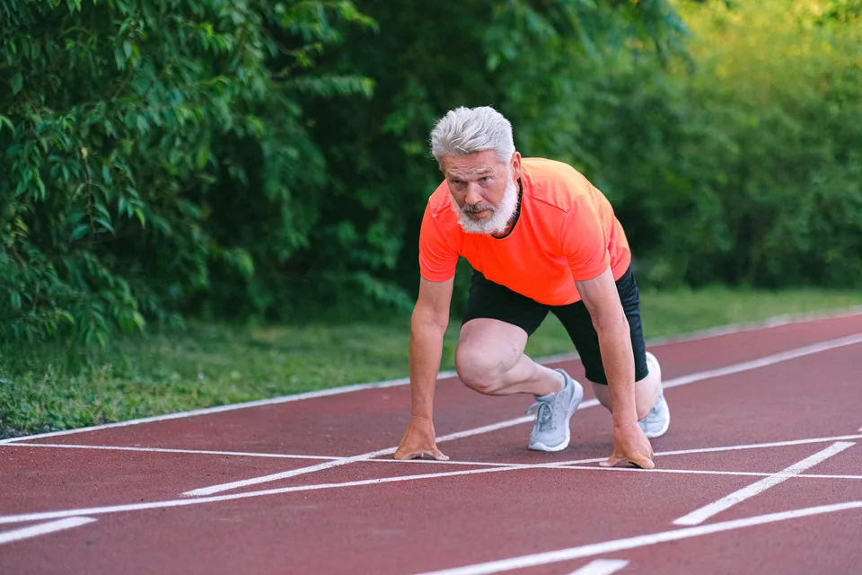 Homem de cabelo branco em posição de largada numa pista de atletismo cercada por árvores