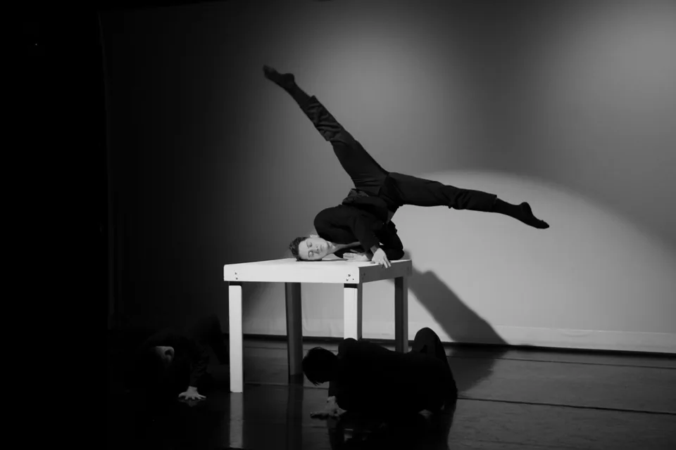 Bailarino em equilíbrio sobre uma mesa branca no palco, em foto em preto e branco, com outros dois dançarinos no chão ao fundo