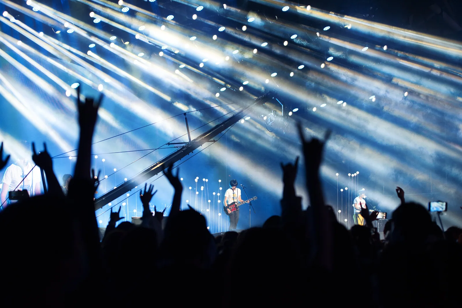 Plateia de silhueta com as mãos levantadas em sinal de rock diante de um palco com feixes de luz azul e um guitarrista ao fundo (foto ilustrativa)