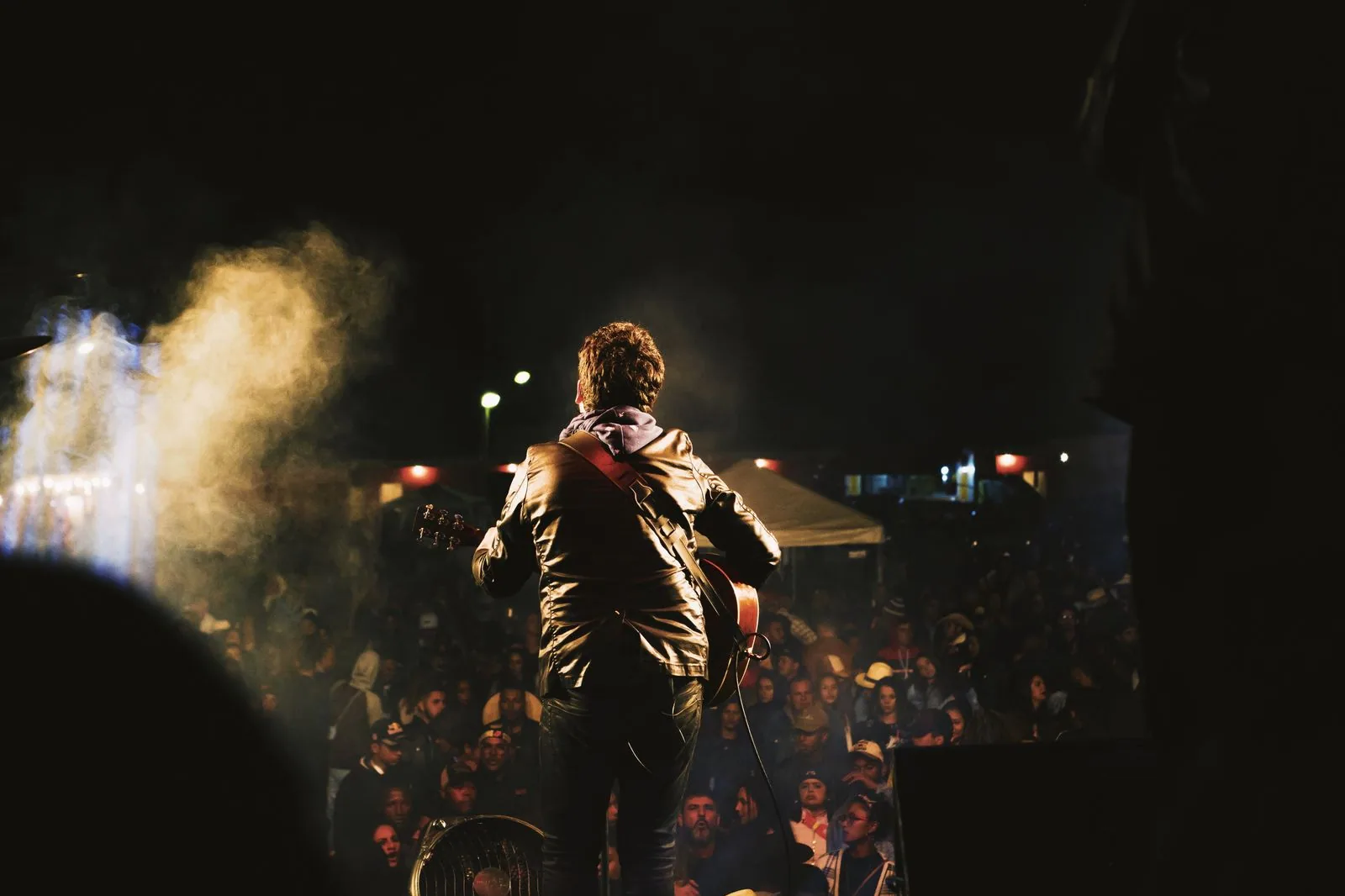 Guitarrista de costas no palco diante de uma plateia à noite, com fumaça e luz amarela (foto ilustrativa)