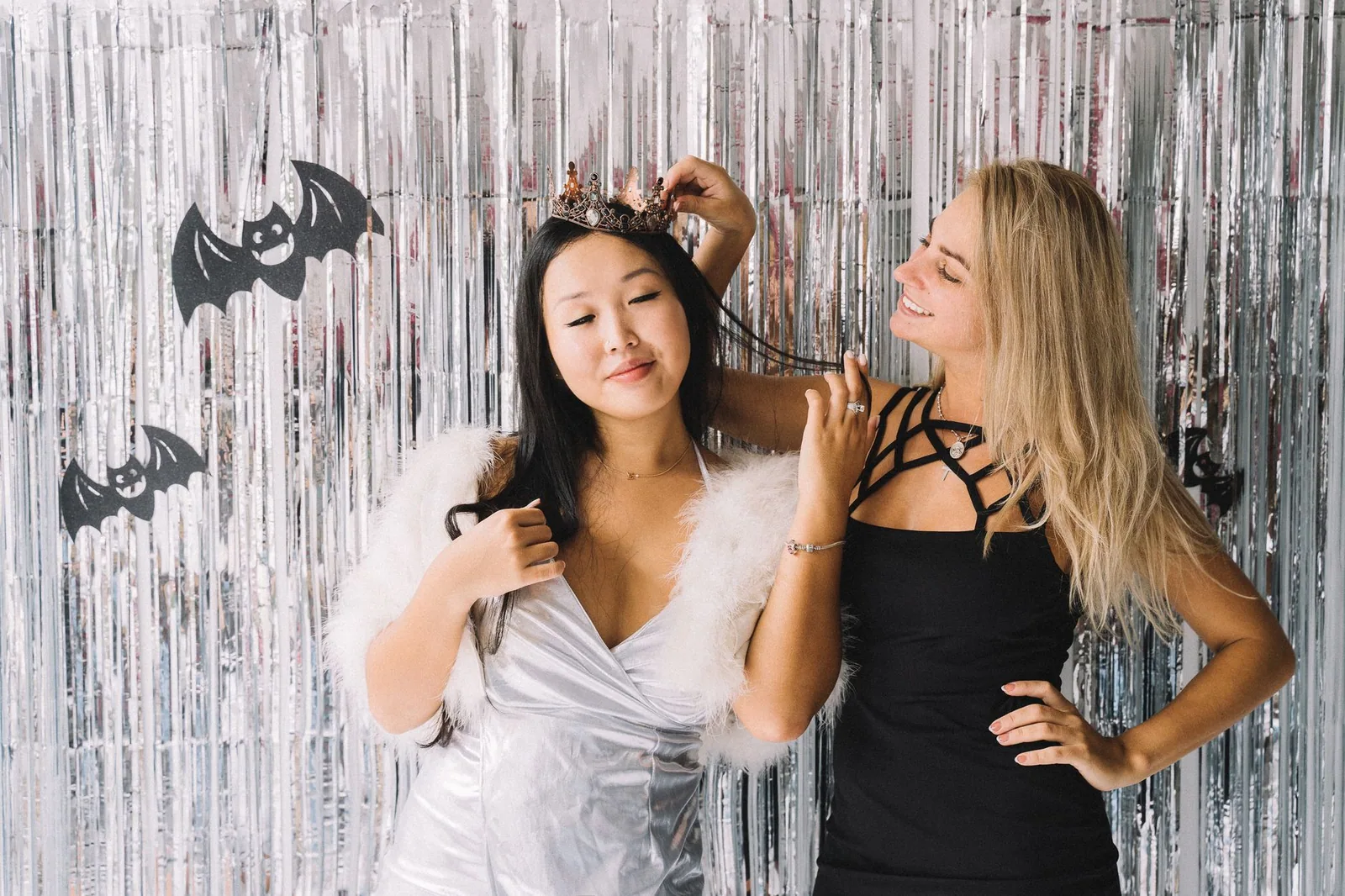 Duas mulheres posam fantasiadas diante de cortina prateada com morcegos de papel, foto ilustrativa de festa de Halloween