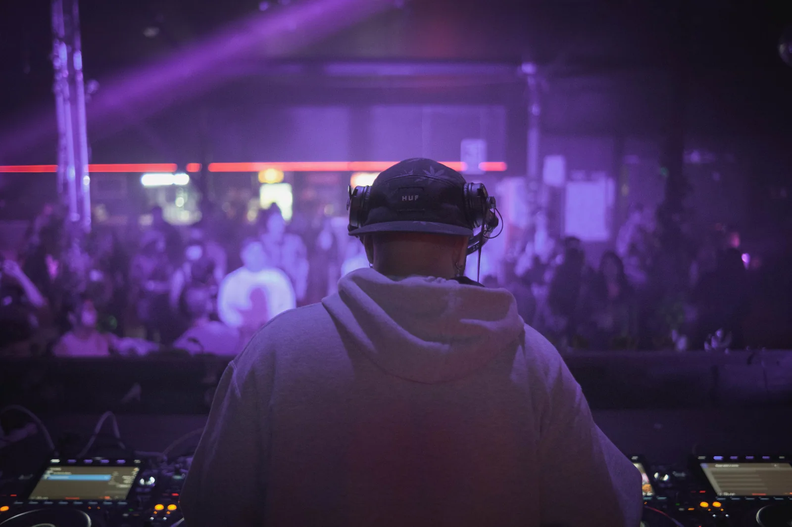 DJ de boné e capuz de costas, com fones no pescoço, toca em pista com luz roxa e público ao fundo, em foto de arquivo.