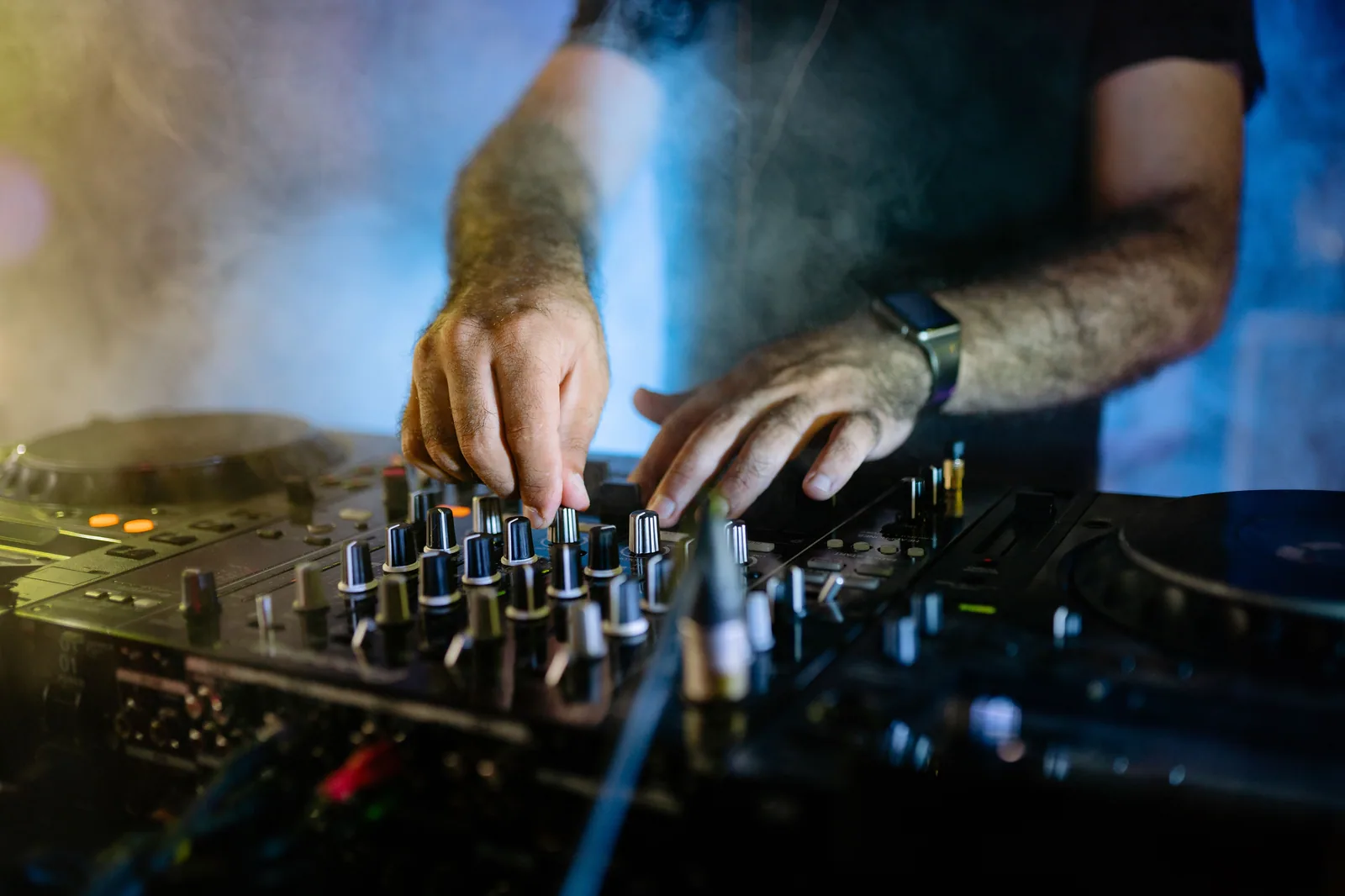 Mãos de DJ sobre a mesa de mixagem, com fumaça e luz azul ao fundo