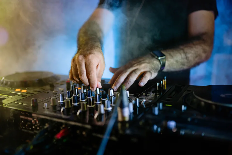 Mãos de DJ sobre a mesa de mixagem, com fumaça e luz azul ao fundo