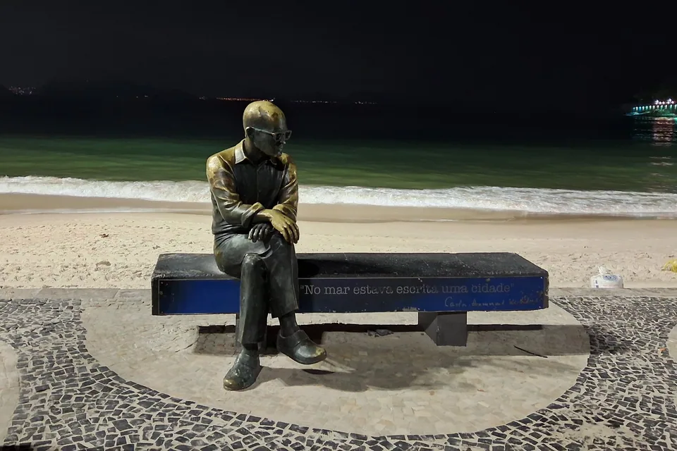Estátua de bronze de Carlos Drummond de Andrade sentado em um banco na orla de Copacabana, com o mar ao fundo
