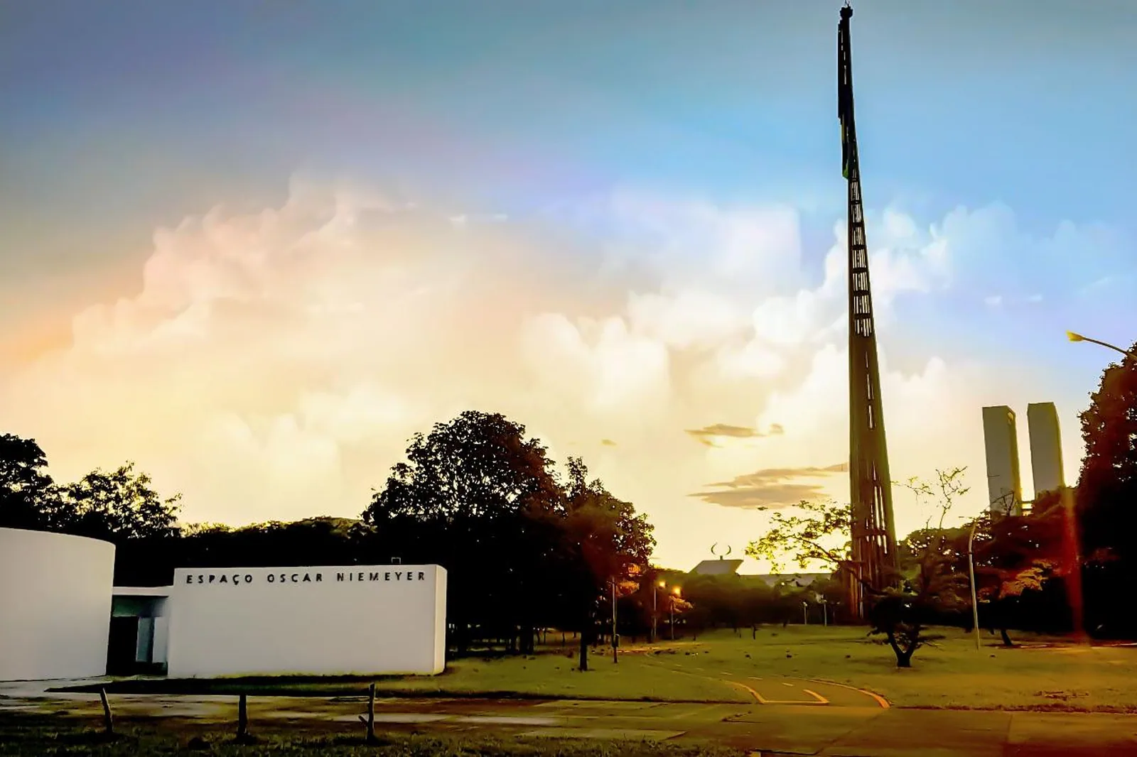 Prédio baixo e branco com a inscrição Espaço Oscar Niemeyer na fachada, gramado à frente, mastro alto e as torres do Congresso ao fundo sob céu de fim de tarde.