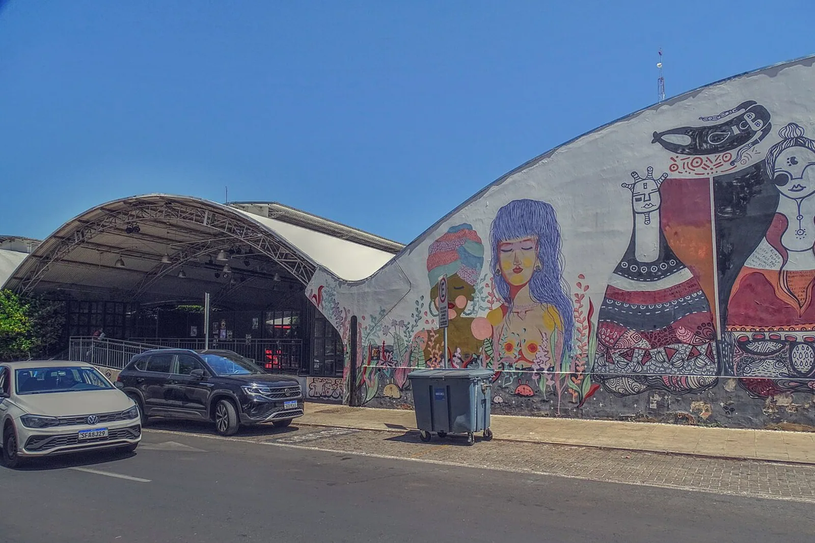 Fachada do Espaço Cultural Renato Russo vista da W2 Sul, com cobertura em arco e muro pintado com mural colorido de figuras femininas, carros estacionados em frente.