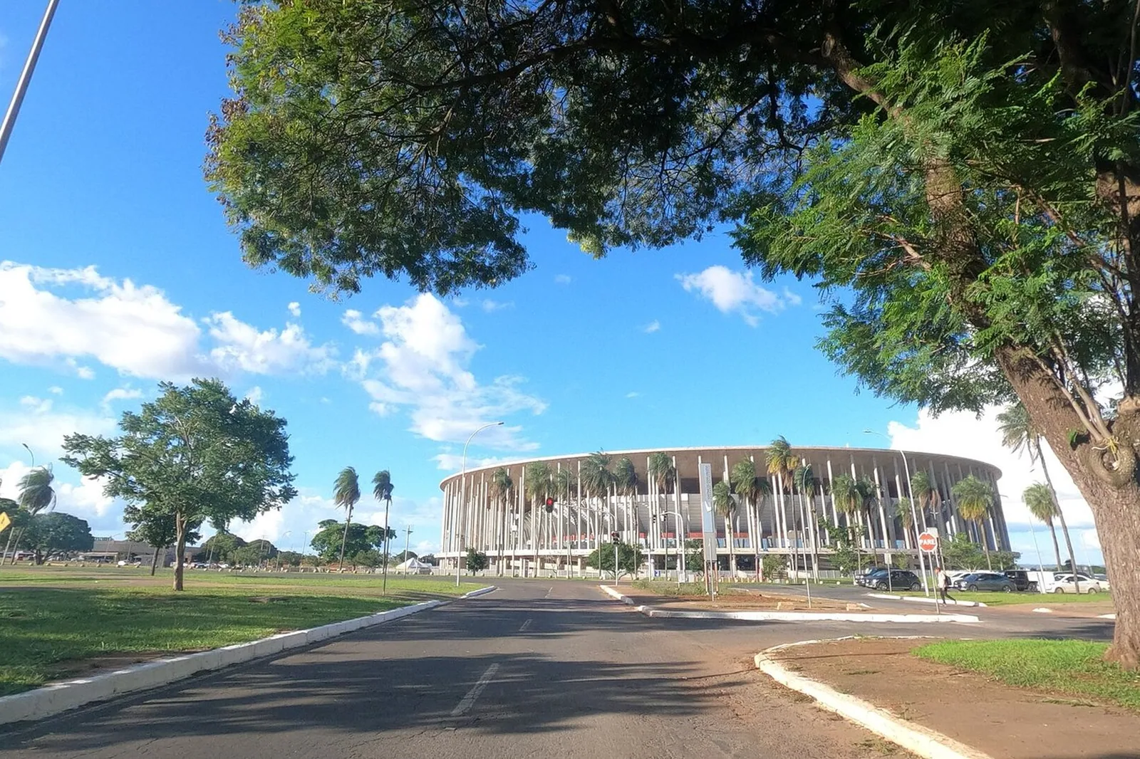 Arena BRB Mané Garrincha vista de uma avenida sombreada por árvores, em dia de céu azul com nuvens