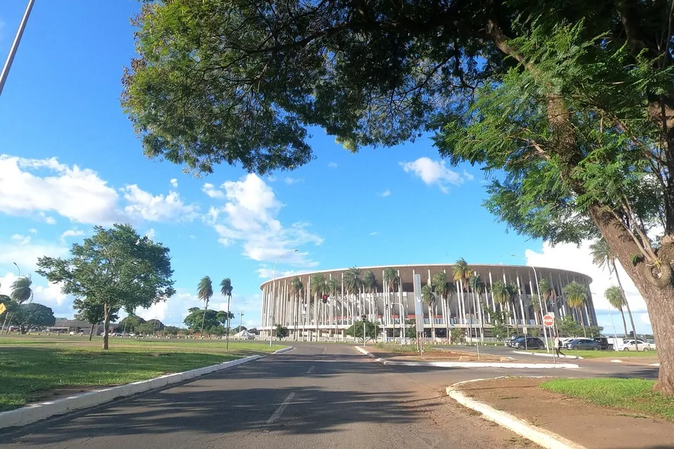 Arena BRB Mané Garrincha vista de uma avenida sombreada por árvores, em dia de céu azul com nuvens