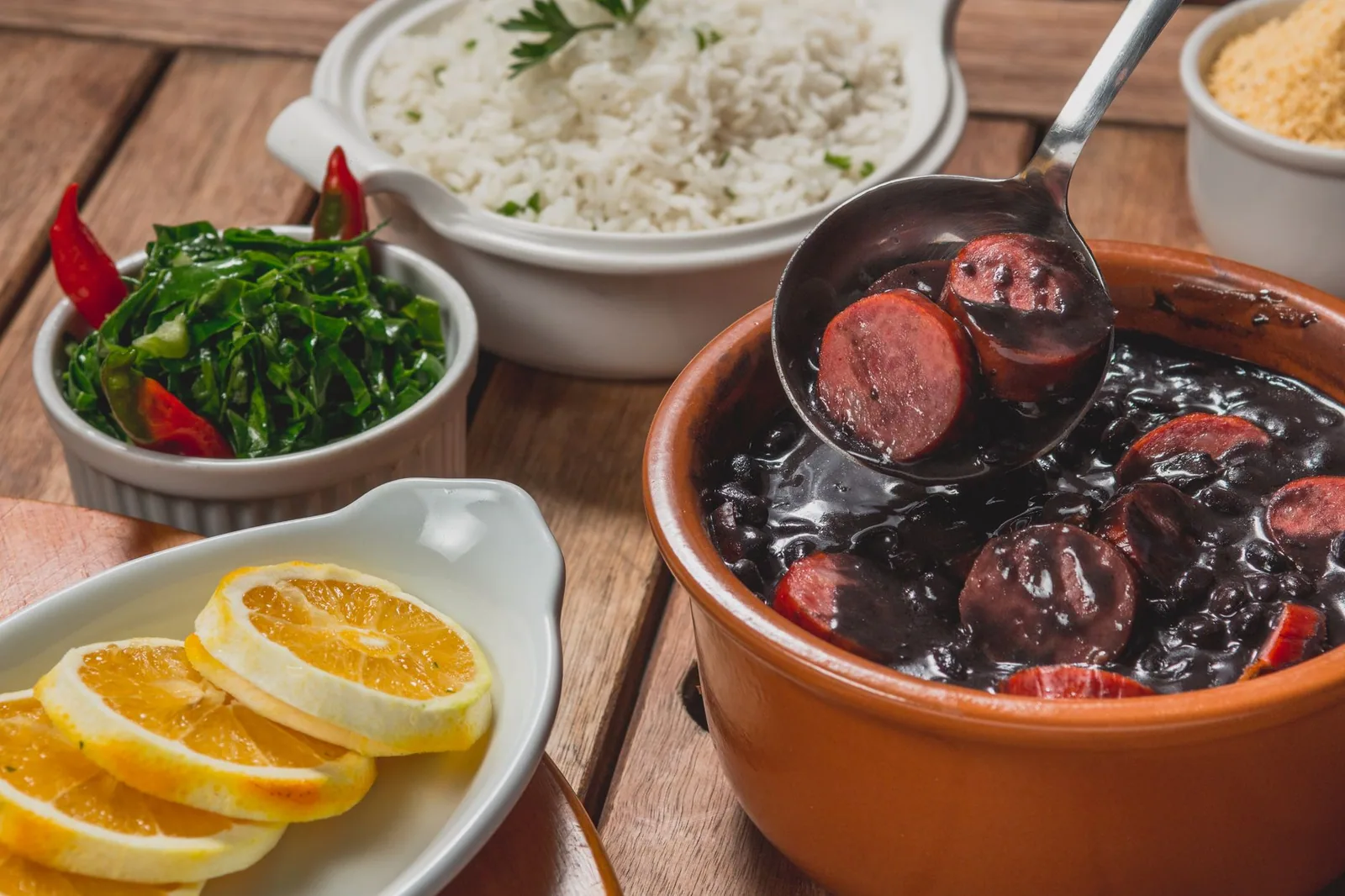 Concha servindo feijoada com linguiça em panela de barro, ao lado de arroz, couve, laranja e farofa