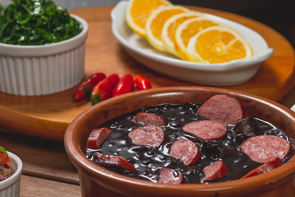 Feijoada servida em panela de barro com rodelas de linguiça, ao lado de couve refogada e fatias de laranja