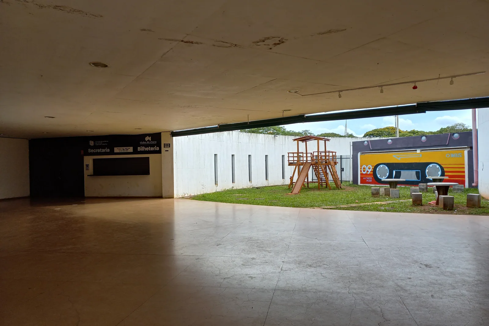 Área coberta do Clube do Choro de Brasília com piso amplo, balcão de bilheteria e parquinho de madeira ao fundo