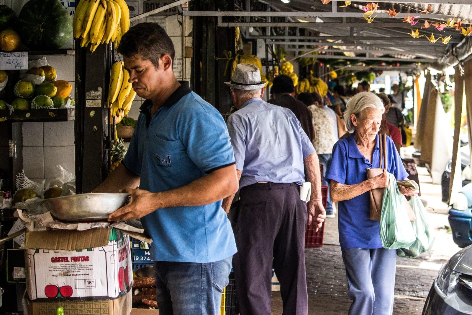 Corredor coberto de feira permanente brasileira, com barracas de frutas e feirantes atendendo clientes.