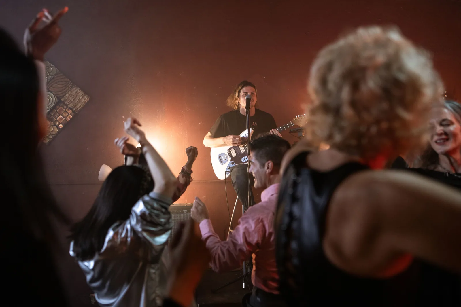 Guitarrista tocando e cantando em um bar, com a plateia de braços levantados em primeiro plano (foto ilustrativa)
