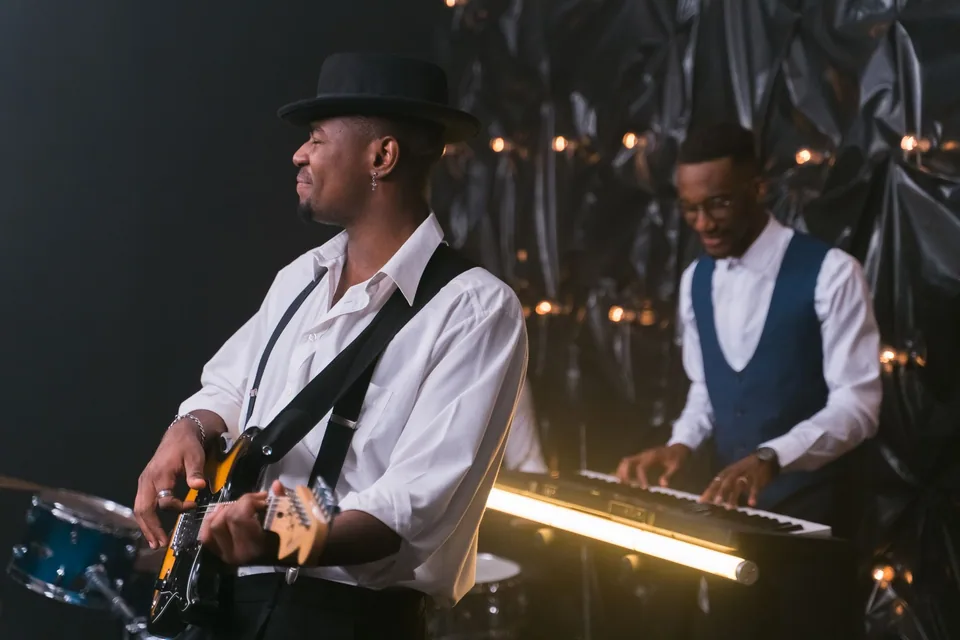 Dois músicos de camisa branca tocando guitarra e teclado em palco escuro com luzes quentes (foto ilustrativa)