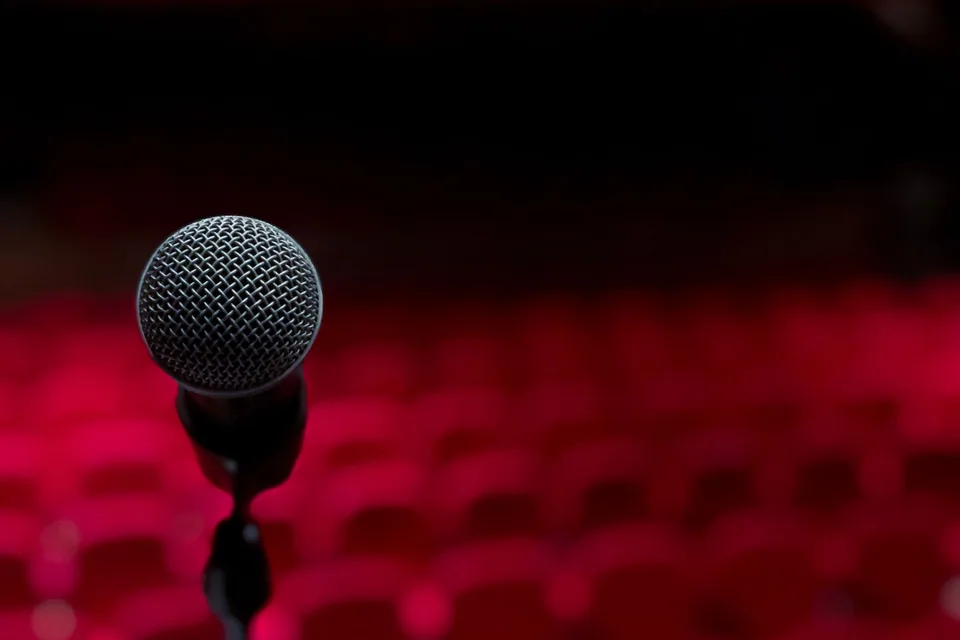 Microfone em close no palco de um teatro, com poltronas vermelhas desfocadas ao fundo (foto de banco de imagens, não é do espetáculo)