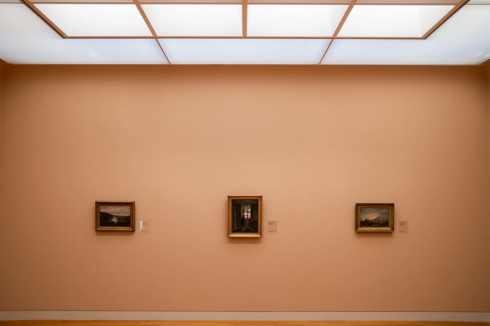 Parede de galeria de arte com três quadros emoldurados e etiquetas ao lado, sob luz difusa de teto