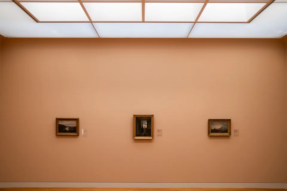 Parede de galeria de arte com três quadros emoldurados e etiquetas ao lado, sob luz difusa de teto