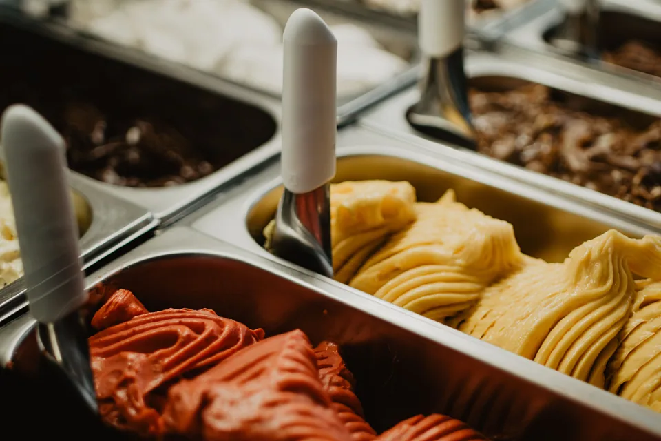 Cubas metálicas de gelateria com sorvete vermelho e amarelo e colheres de servir espetadas