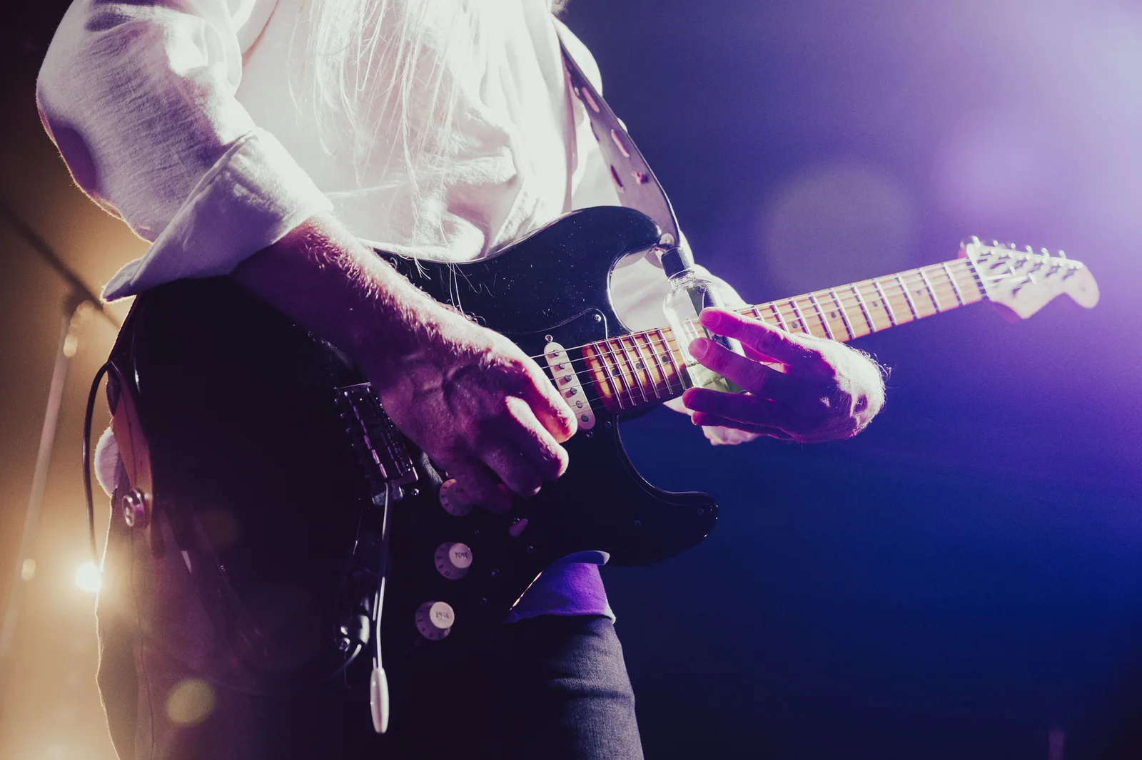 Guitarrista tocando guitarra elétrica preta no palco, sob luz roxa e dourada