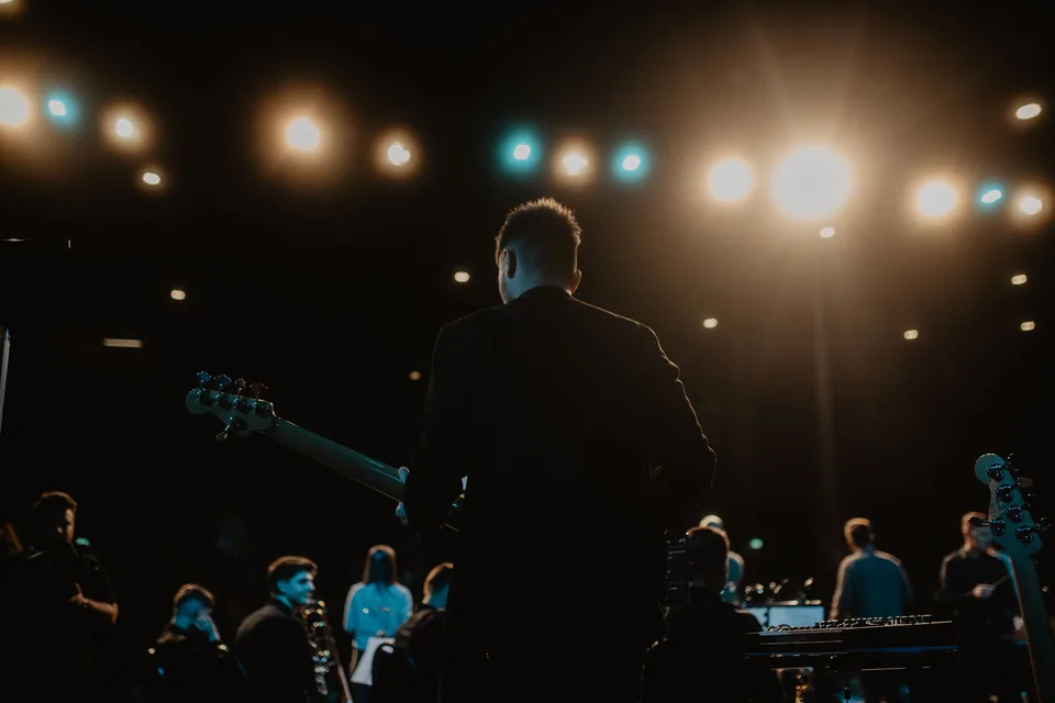 Guitarrista de costas no palco, sob luzes quentes e azuis, com a banda e o público ao fundo