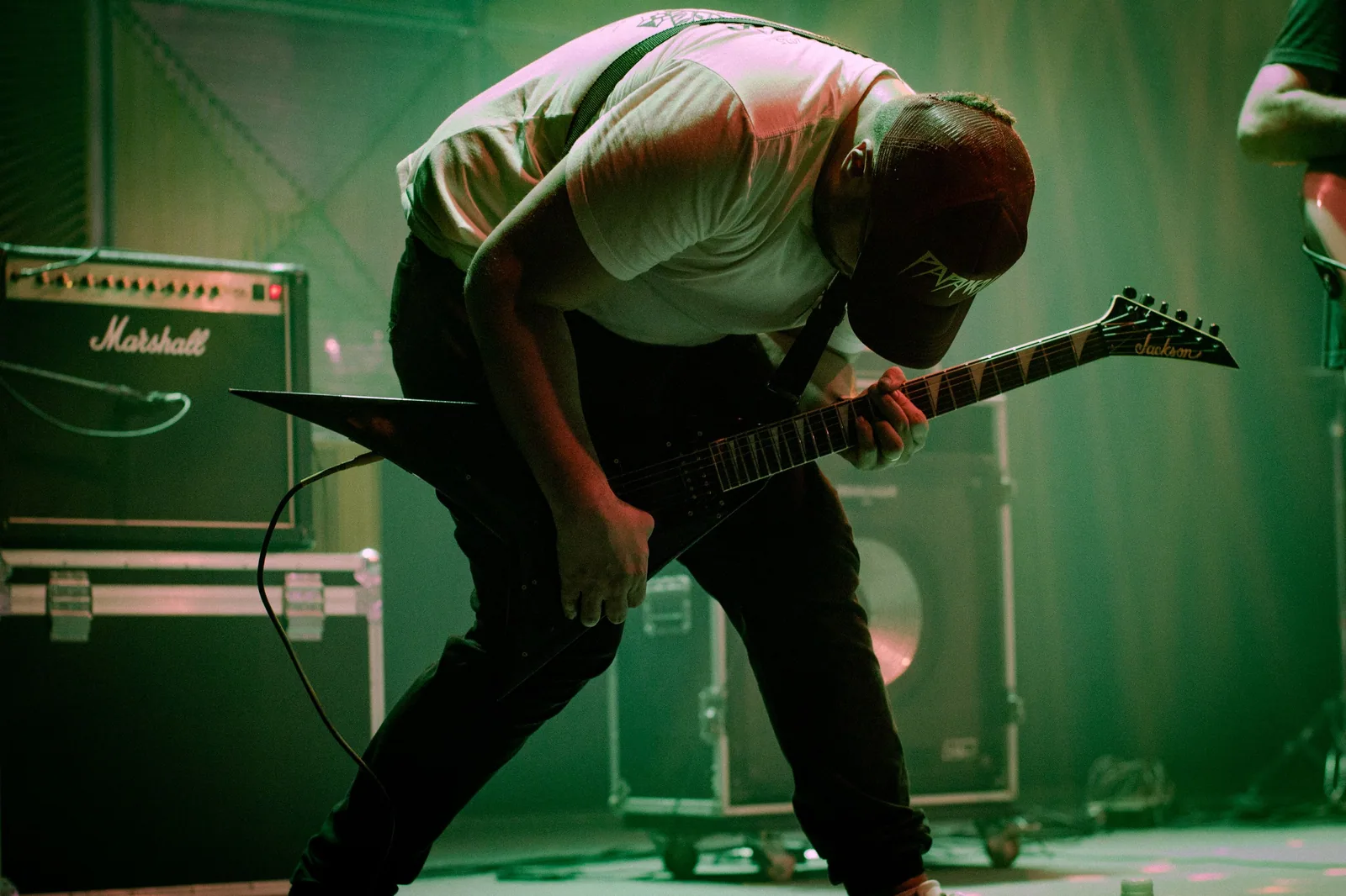 Guitarrista curvado sobre uma guitarra de corpo em V diante de um amplificador, sob luz verde de palco