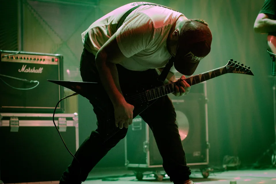 Guitarrista curvado sobre uma guitarra de corpo em V diante de um amplificador, sob luz verde de palco