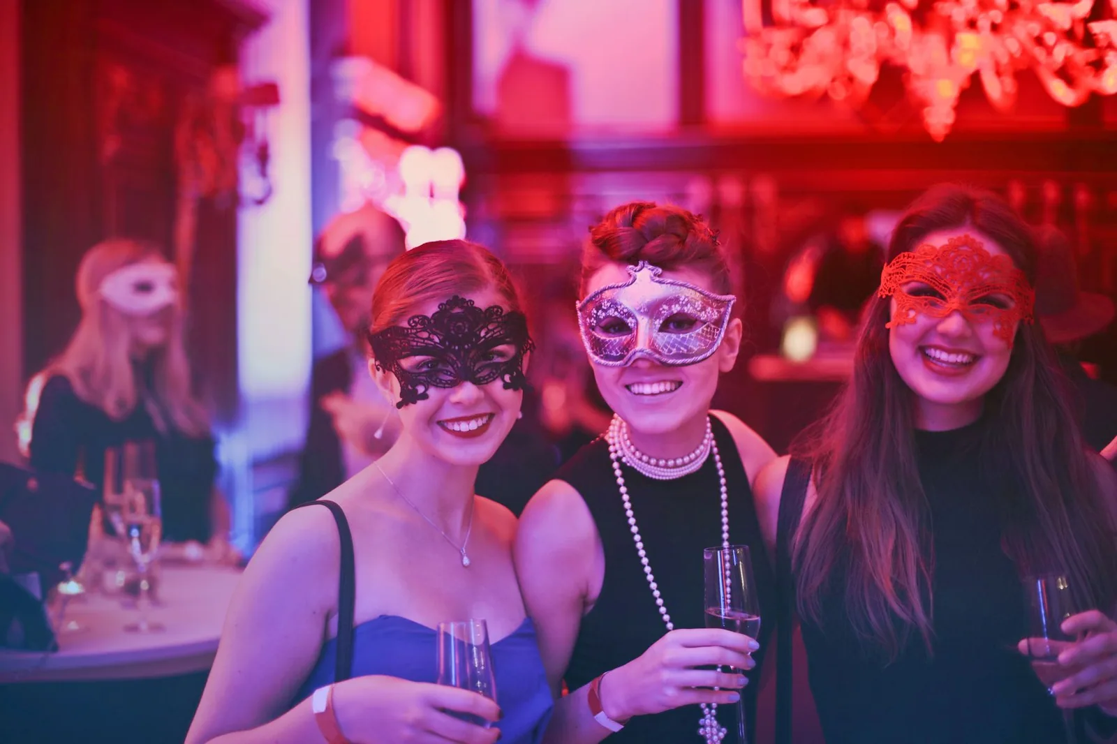Três mulheres de máscara de baile, uma preta, uma prateada e uma vermelha, seguram taças em uma festa com luz rosa e vermelha