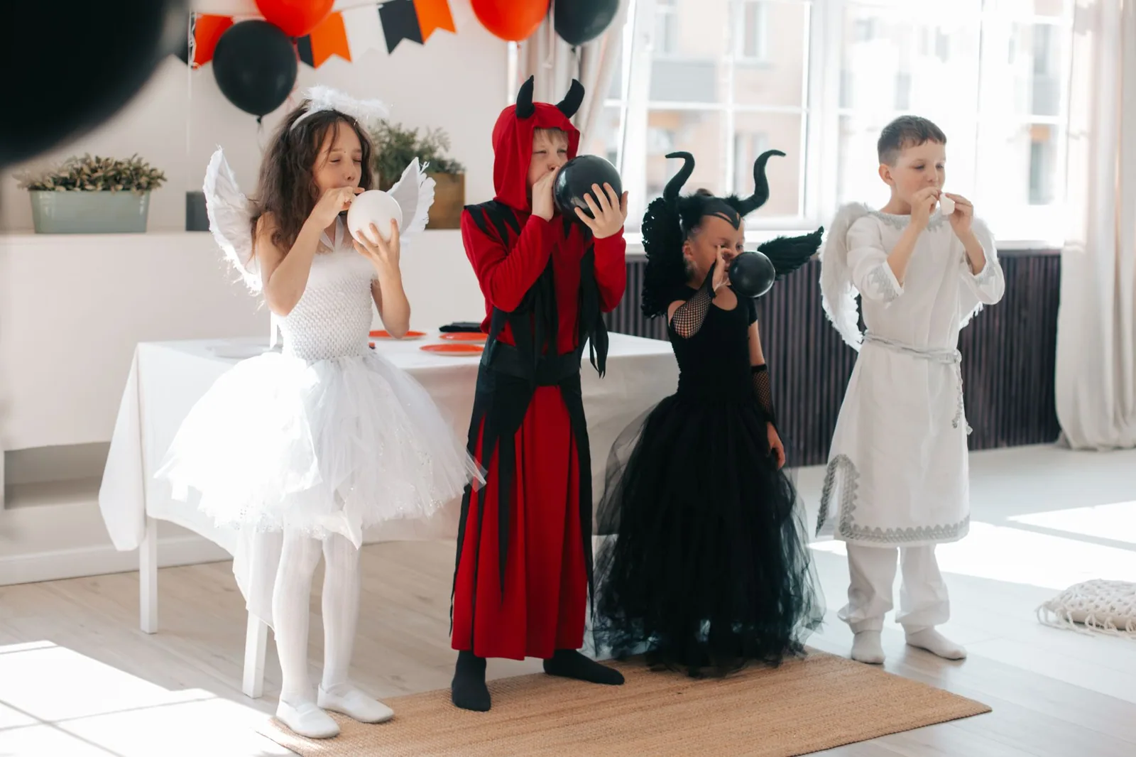 Quatro crianças fantasiadas de anjo e de diabo enchem balões em uma festa de Halloween