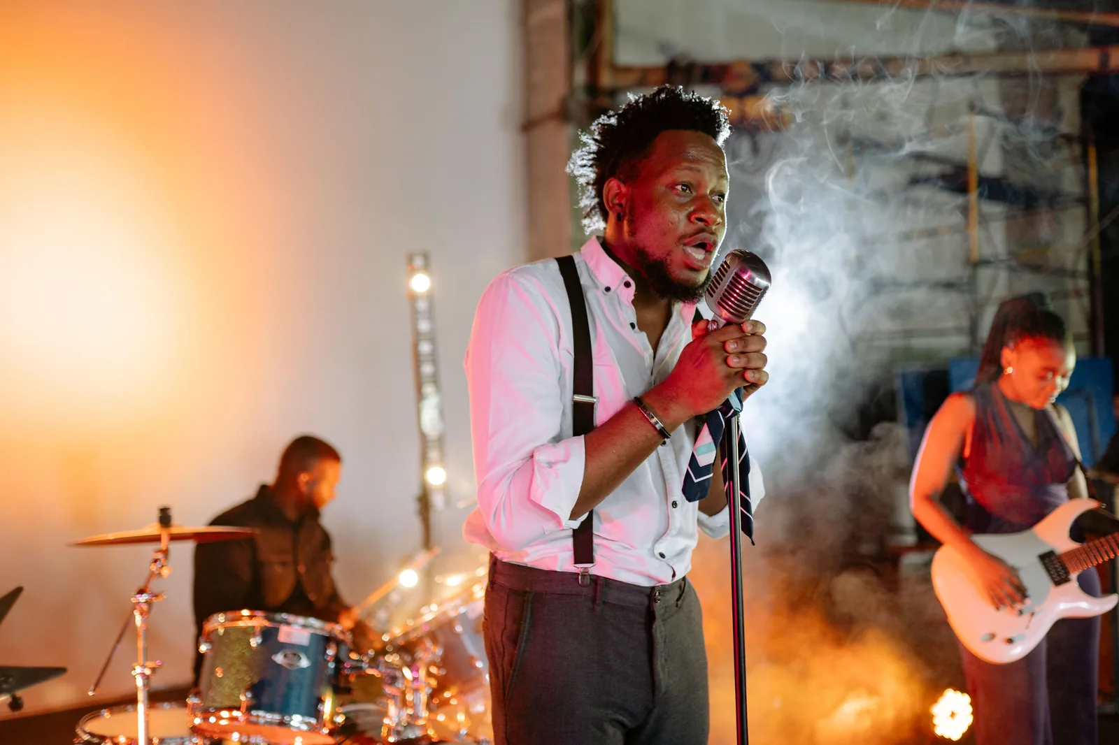 Cantor ao microfone com baterista e guitarrista em palco com fumaça, foto ilustrativa