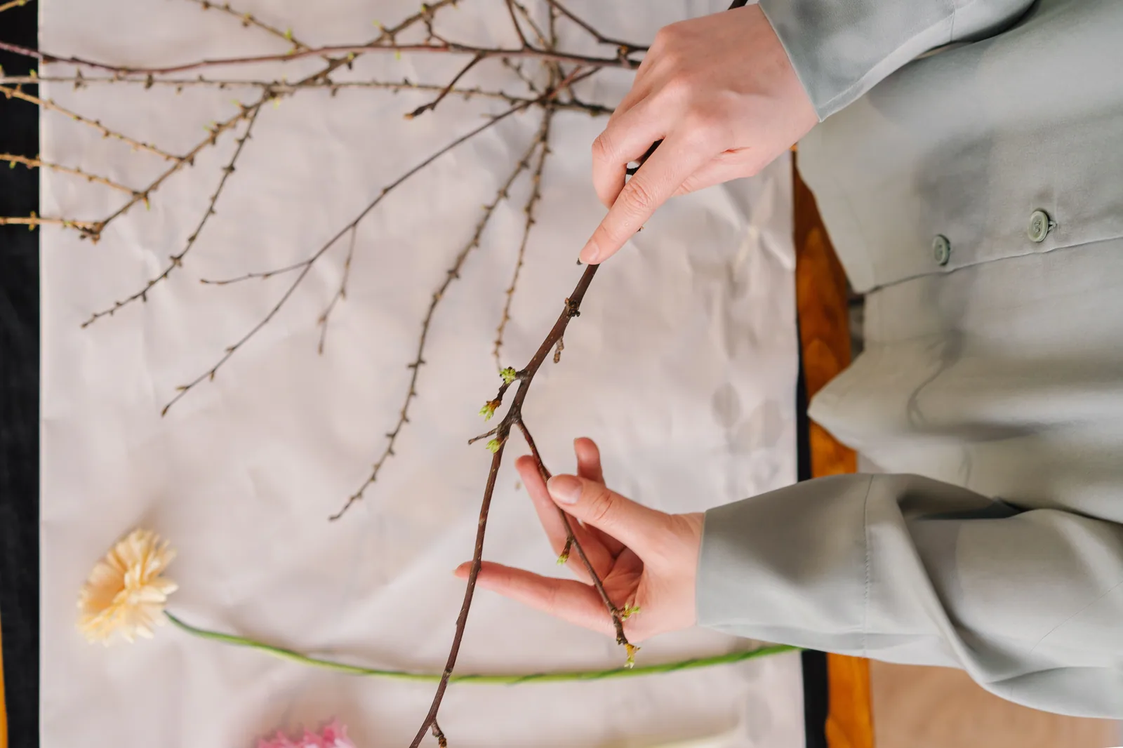 Mãos arrumam galhos e flores sobre uma mesa, em preparo de arranjo no estilo ikebana