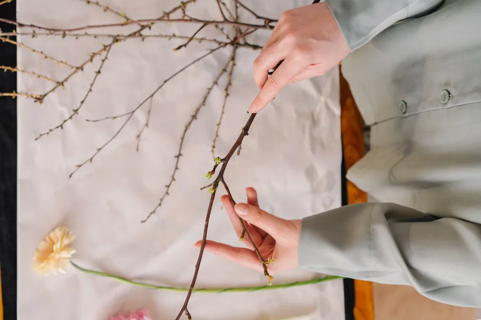 Mãos arrumam galhos e flores sobre uma mesa, em preparo de arranjo no estilo ikebana
