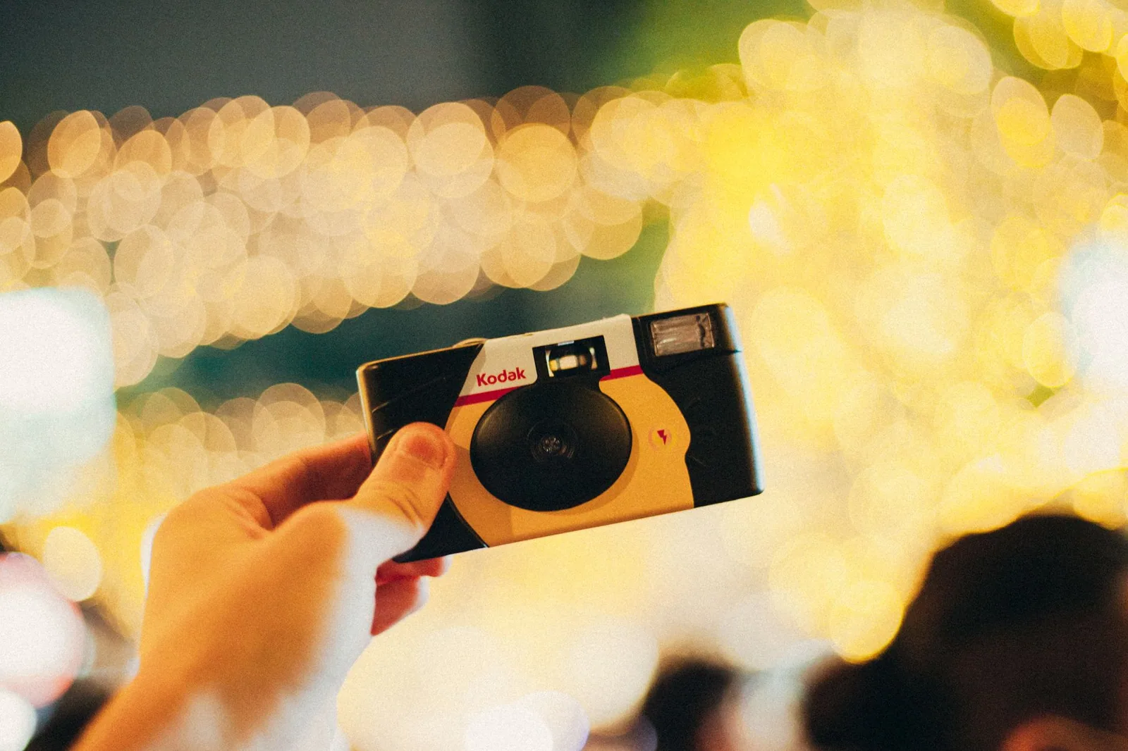 Mão segura uma câmera descartável amarela e preta diante de luzes desfocadas, em foto ilustrativa que lembra o visual fotolog da festa