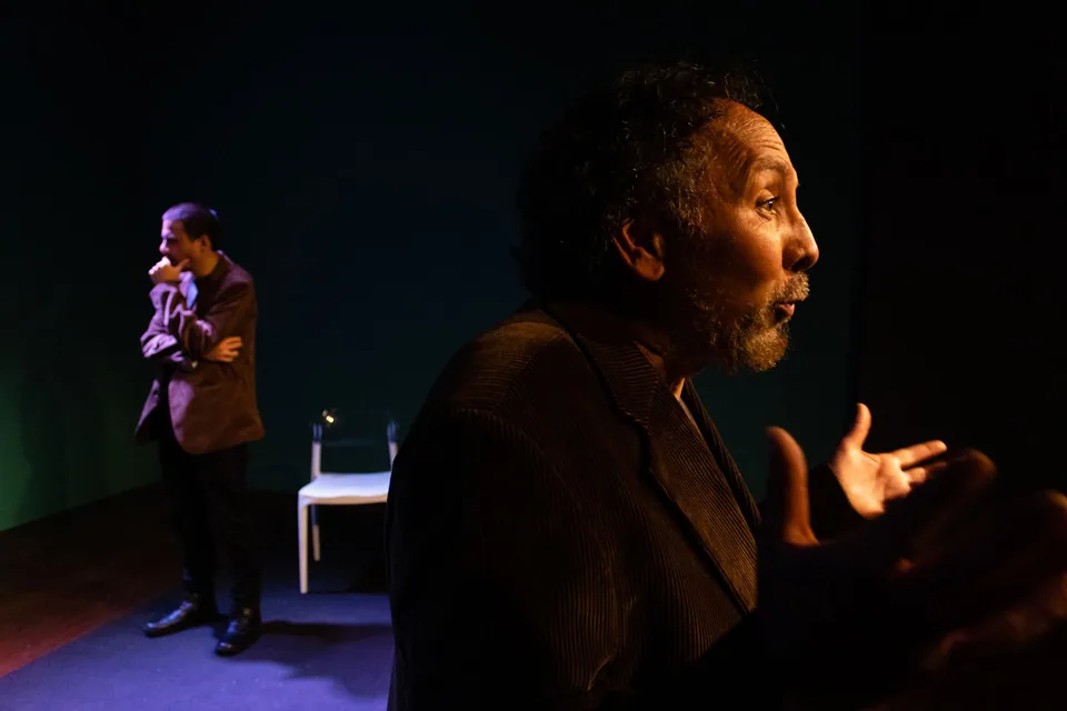 Dois atores em cena em palco escuro, um em primeiro plano gesticulando e outro ao fundo com a mão no queixo, ao lado de uma cadeira branca
