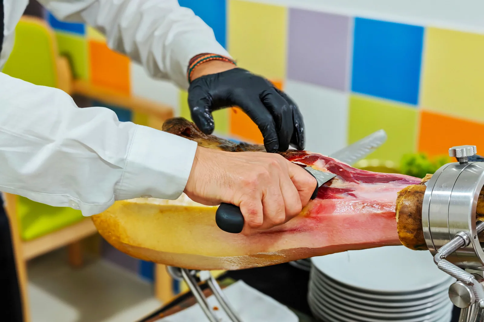Mãos de profissional cortando fatia de jamón preso em uma jamonera, com pratos empilhados ao lado