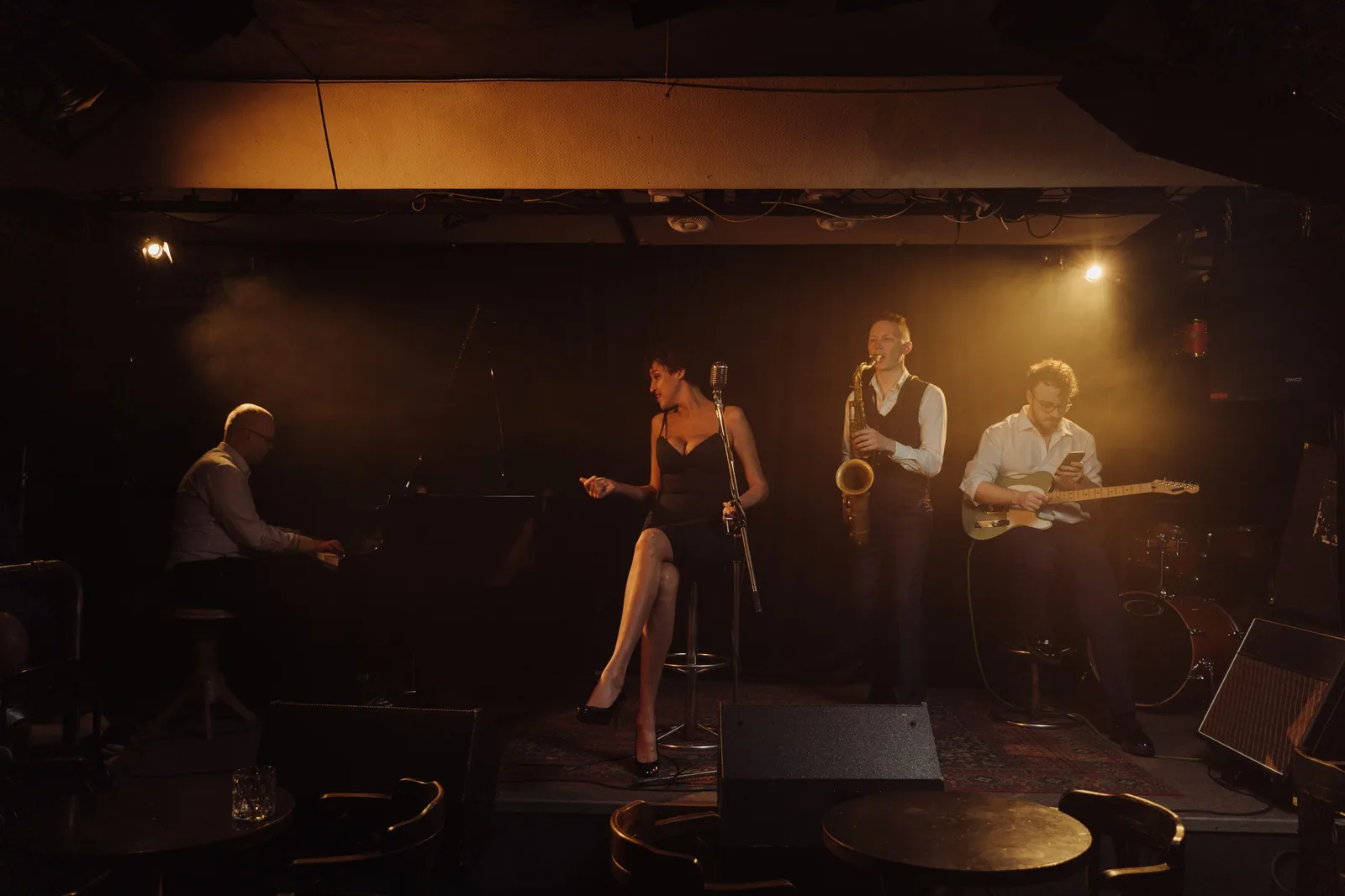 Banda de jazz tocando em palco de clube com luz quente, com piano, saxofone, guitarra e vocalista