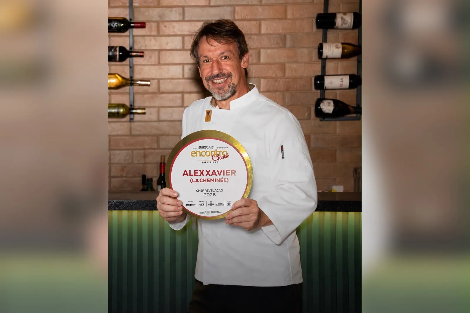 O chef Alex Xavier, de dólmã branca, sorri segurando a placa de Chef Revelação 2026 do prêmio Encontro Gastrô, diante de uma parede de tijolos com garrafas de vinho