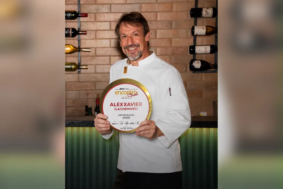 O chef Alex Xavier, de dólmã branca, sorri segurando a placa de Chef Revelação 2026 do prêmio Encontro Gastrô, diante de uma parede de tijolos com garrafas de vinho