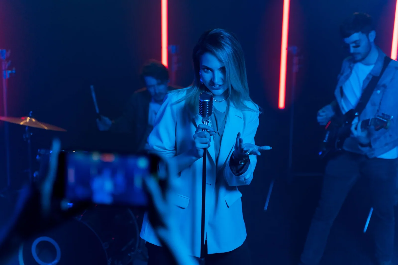 Cantora com microfone de pedestal em palco com luz azul e neon vermelho, com baterista e guitarrista ao fundo