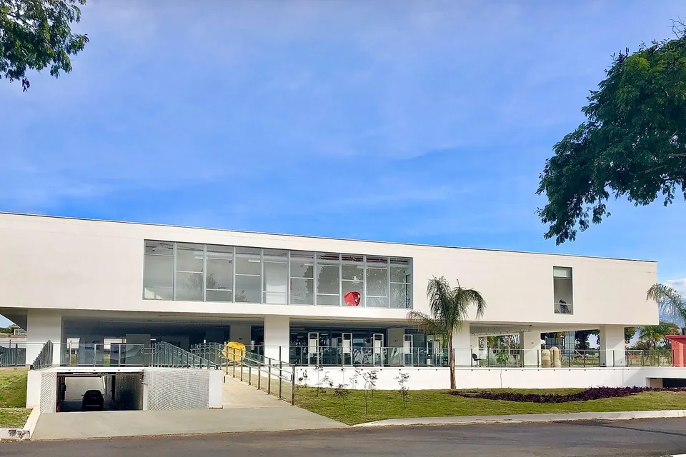 Prédio branco de dois andares do Museu de Arte de Brasília, com faixa de vidro no pavimento superior, gramado e árvores na frente sob céu azul.