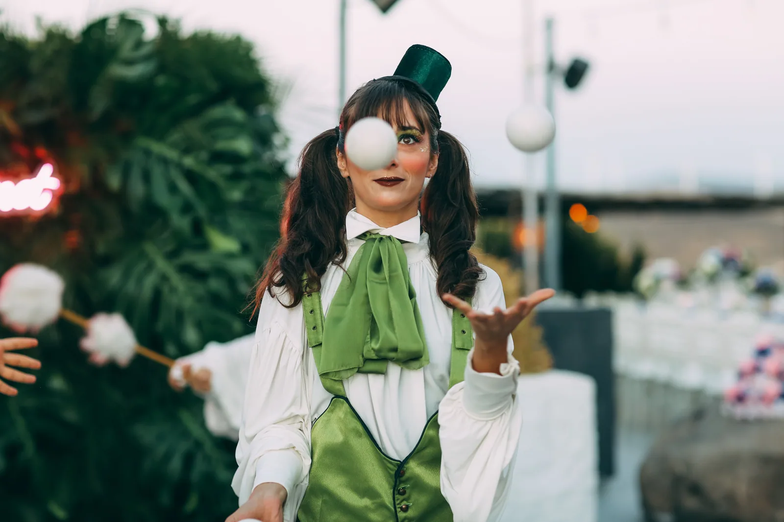 Artista circense de colete verde e cartola faz malabarismo com bolas brancas ao ar livre