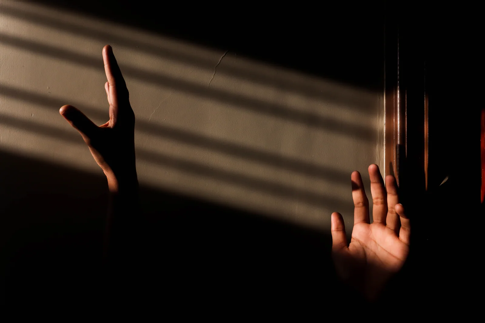 Duas mãos erguidas diante de uma parede com faixas de luz e sombra, foto ilustrativa que não é do espetáculo