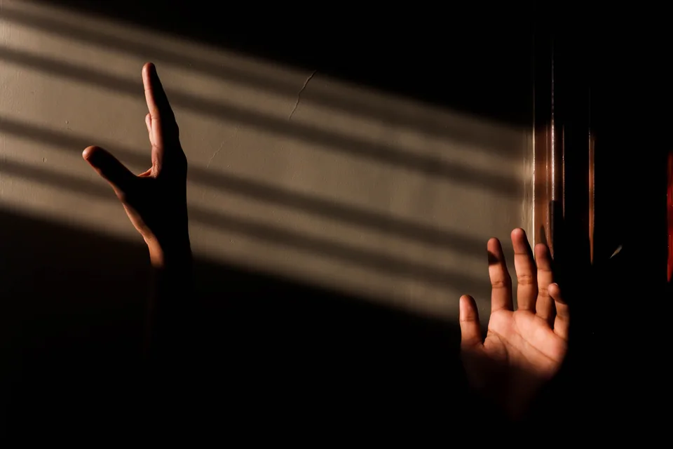 Duas mãos erguidas diante de uma parede com faixas de luz e sombra, foto ilustrativa que não é do espetáculo
