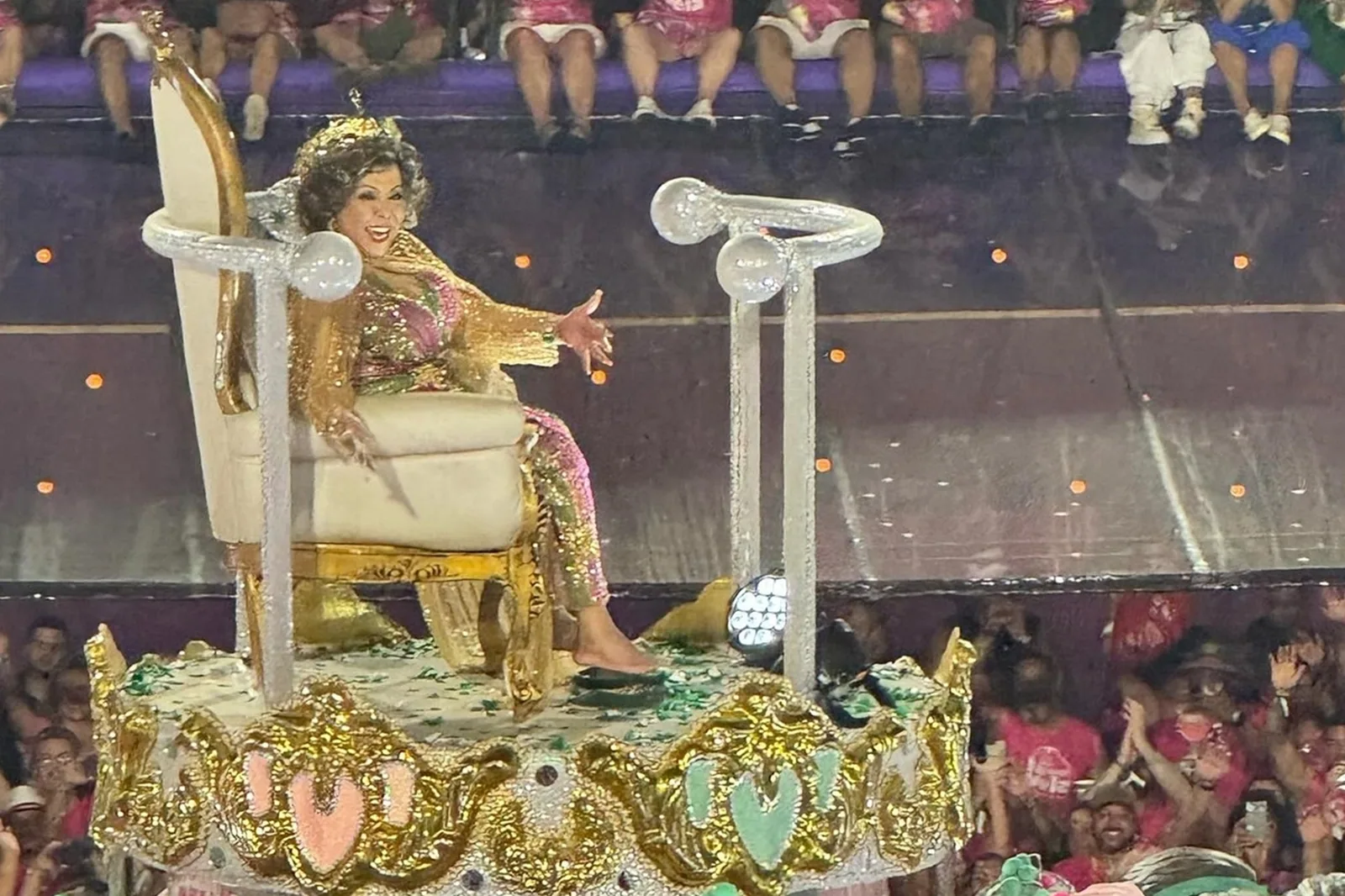 Alcione sentada em um trono dourado sobre um carro alegórico, de braços abertos, em desfile de carnaval