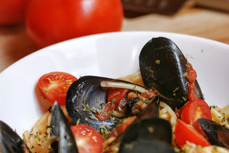Massa com mexilhões, tomate-cereja e azeitonas pretas em prato branco fundo