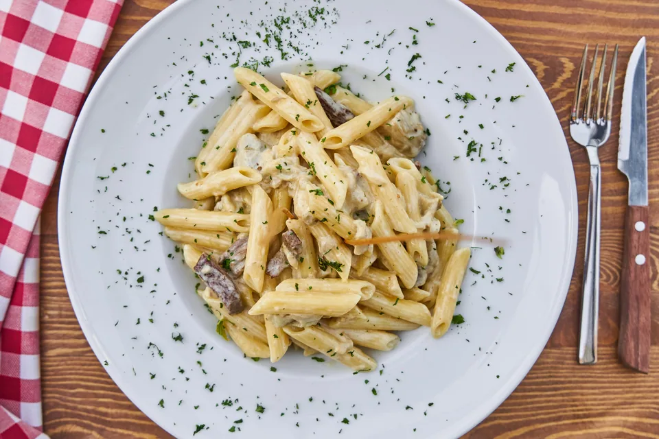 Prato branco com penne ao molho cremoso sobre mesa de madeira, ao lado de garfo e faca