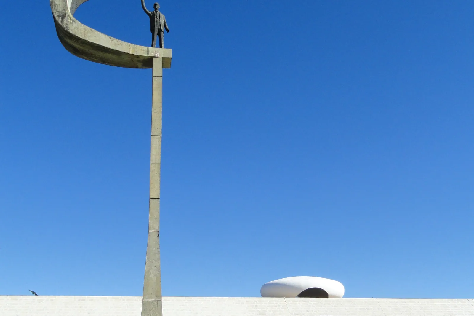 Estátua de Juscelino Kubitschek acenando do alto do Memorial JK, com o Panteão da Pátria ao fundo