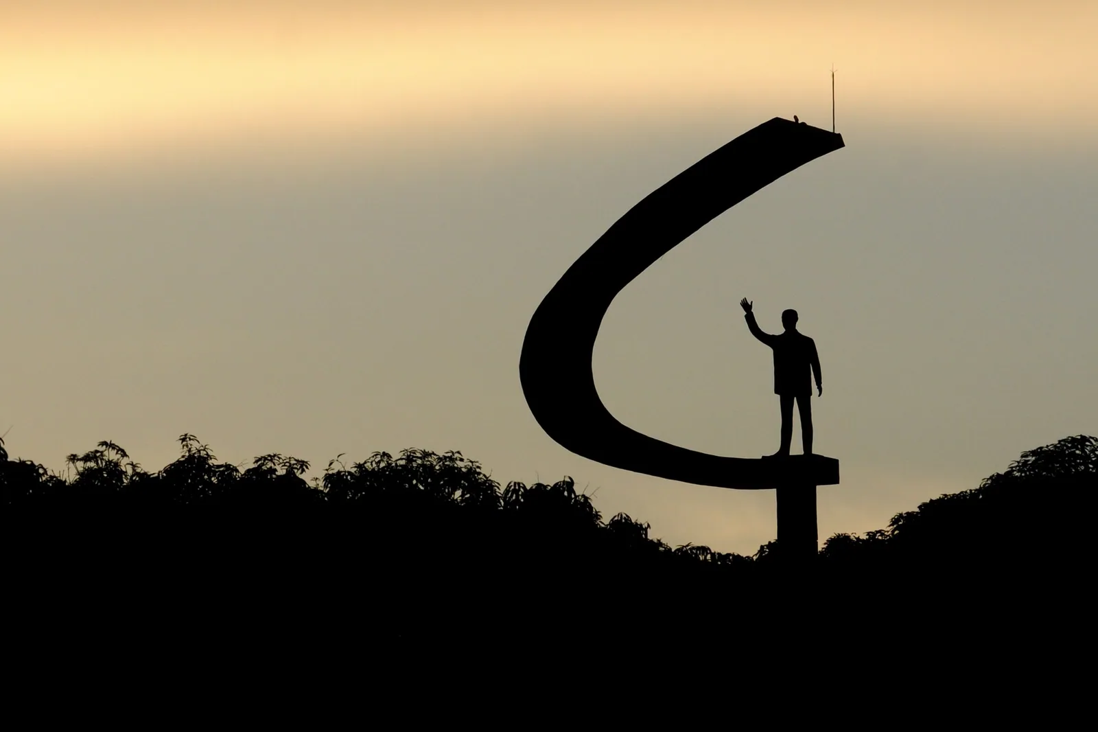 Silhueta do Memorial JK e da estátua de Juscelino Kubitschek com a mão erguida, contra o céu do fim de tarde.