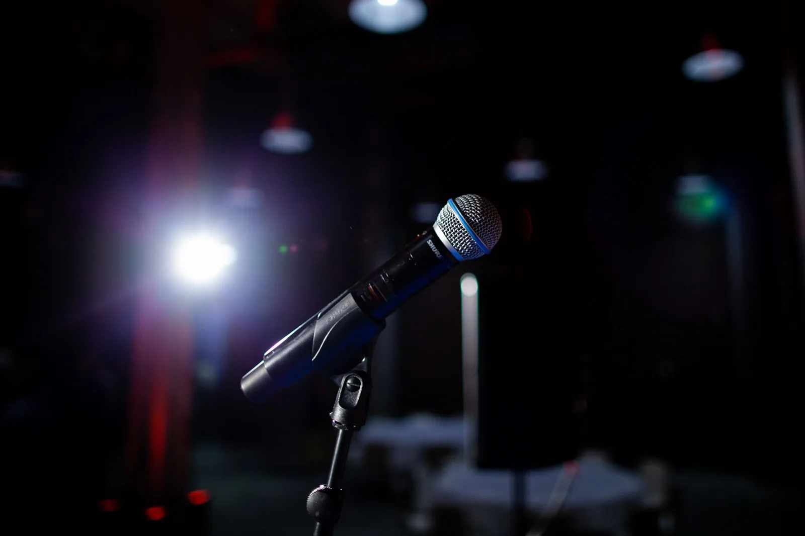 Microfone em pedestal no palco escuro, iluminado por um refletor azul, com luzes desfocadas ao fundo