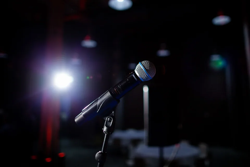 Microfone em pedestal no palco escuro, iluminado por um refletor azul, com luzes desfocadas ao fundo